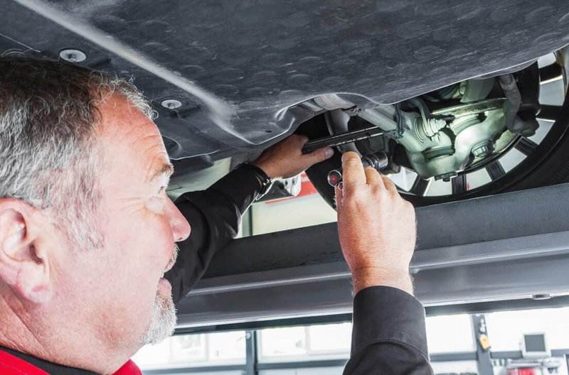 Prüfung Antriebsgelenk Ein Mitarbeiter von GTÜ Neidhardt überprüft mit einem Taschenlampenlicht und Werkzeug das Antriebsgelenk eines Fahrzeugs in einer Werkstatt.