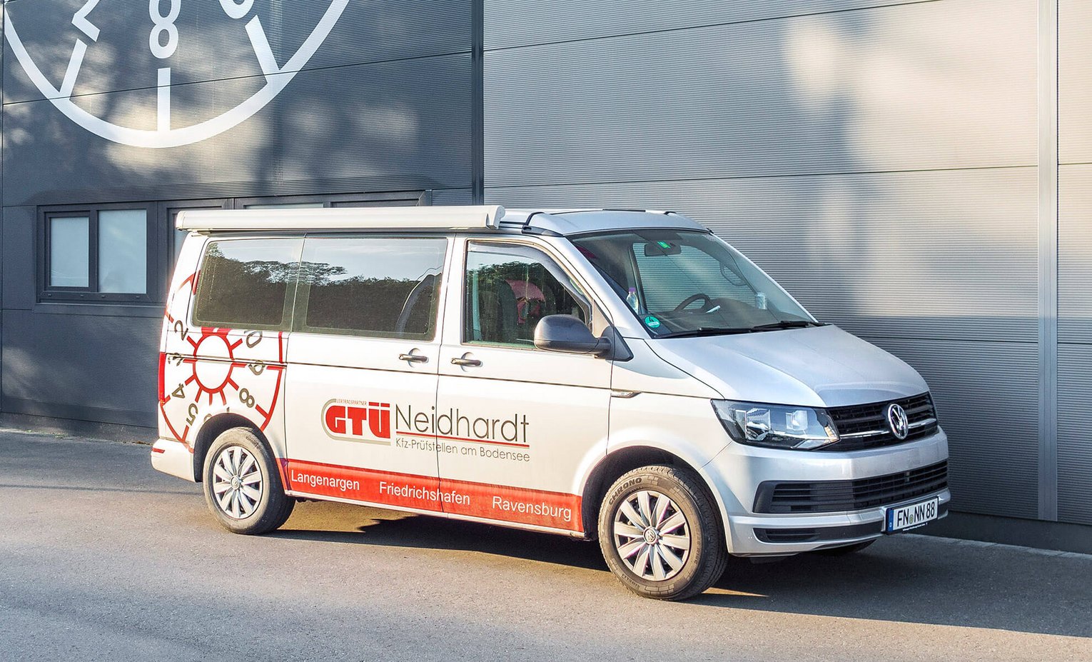 Campingbus vor Gebäude Weißer Volkswagen Transporter mit Werbung für GTÜ Neidhardt in Friedrichshafen, Überlingen und Ravensburg, geparkt vor einer grauen Wand..