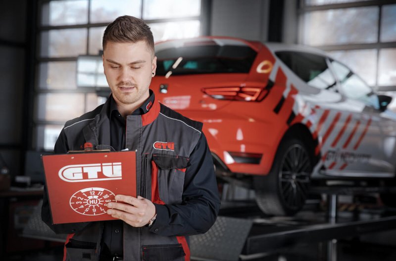 Kfz-Sachverständige GTÜ Neidhardt Ein Mitarbeiter von GTÜ Neidhardt überprüft eine Prüfliste auf einem Klemmbrett, im Hintergrund steht ein rotes Auto auf einer Hebebühne.